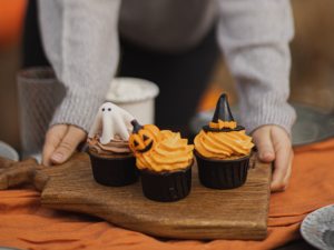 Imagem ilustrativa de três cupcakes de chocolate em cima de uma tábua. O da direita tem topo de fantasma, o do meio é uma abóbora laranja de chantilly e, o terceiro, tem o mesmo chantilly laraja, com um chapéu preto de bruxa no topo.