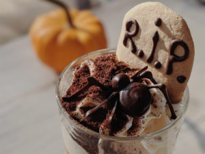 Foto ilustrativa de um copo de vidro com sorvete de chocolate. No topo, uma aranha feita de chocolate e uma lápide de bolacha, escrito RIP em chocolate. Ao fundo, desfocado, tem uma abóbora laranja.