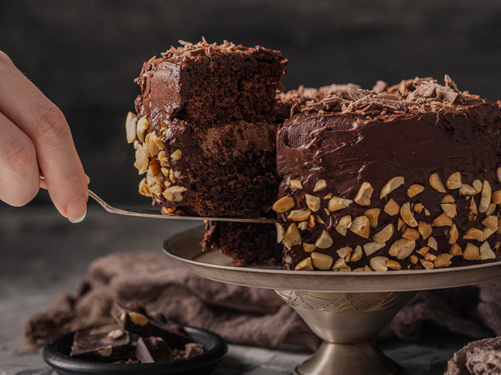 Fotografia ilustrativa de um bolo de chocolate e amêndoas de tamanho médio. Na direita, já uma mão retirando um pedaço com uma espátula. Conteúdo em Confeitaria lucrativa: esteja preparado para inovar